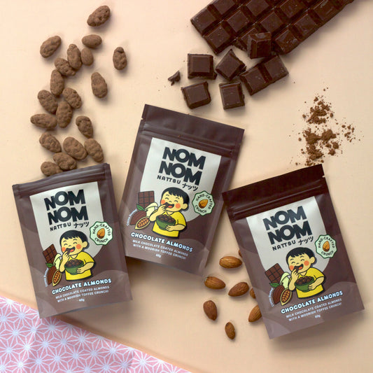 Three packages of 'NOM NOM' chocolate almonds with chocolate pieces and almonds on a light pink background.