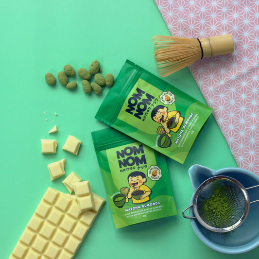 Green packages of 'NOM NOM' matcha almonds on a green background with white chocolate and matcha powder.