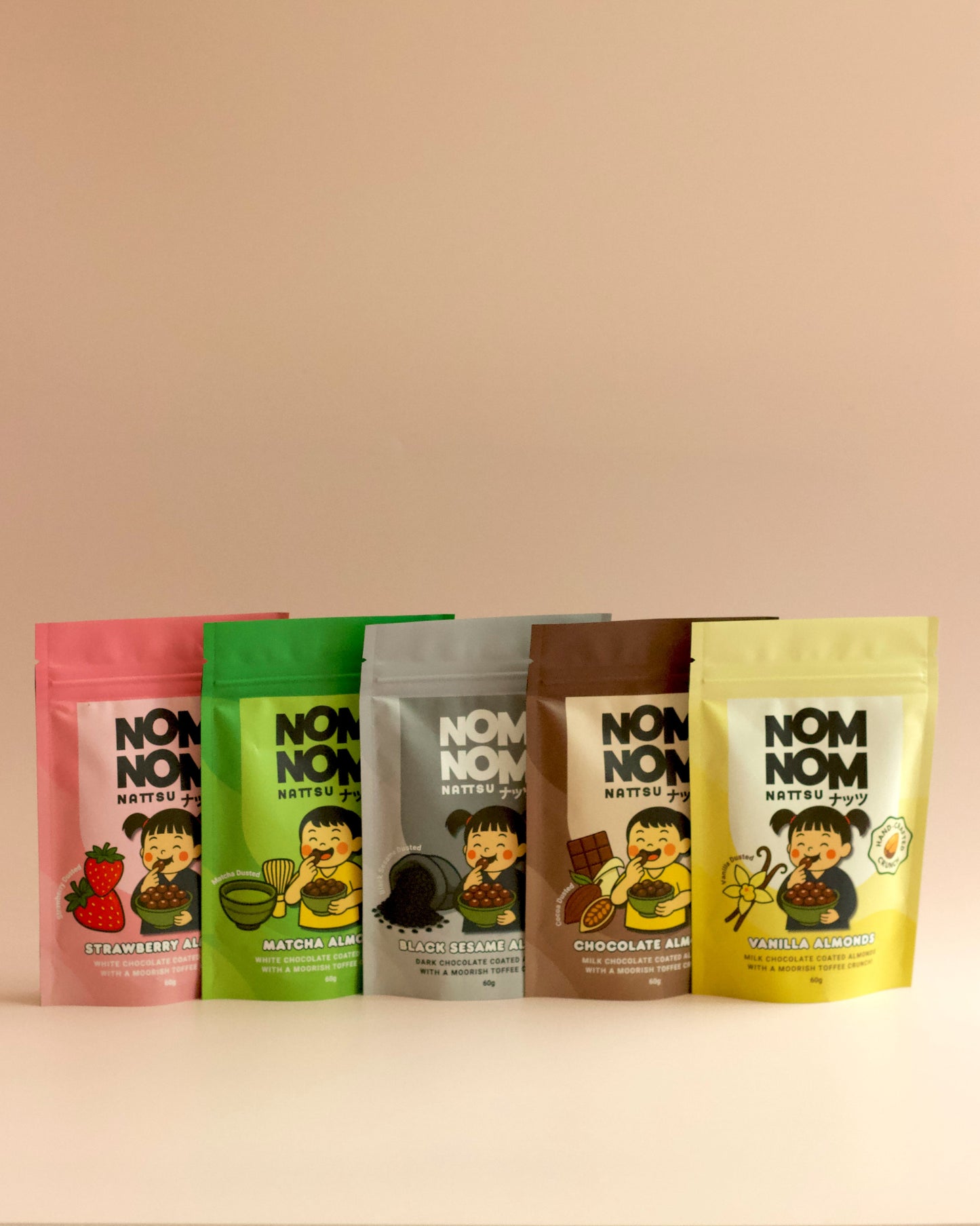 5 NOM NOM 60G POUCHES FULL OF CHOCOLATE COATED ALMONDS IN FLAVOURS - STRAWBERRY, MATCHA, BLACK SESAME, CHOCOLATE AND VANILLA. ALL SET AGAINS A DUSTY PINK BACKGROUND