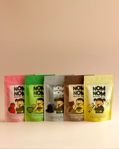 5 NOM NOM 60G POUCHES FULL OF CHOCOLATE COATED ALMONDS IN FLAVOURS - STRAWBERRY, MATCHA, BLACK SESAME, CHOCOLATE AND VANILLA. ALL SET AGAINS A DUSTY PINK BACKGROUND