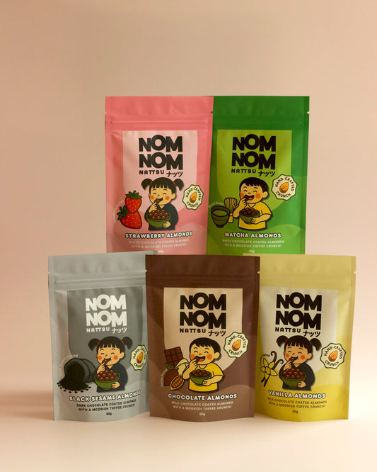 5 NOM NOM 60G POUCHES FULL OF CHOCOLATE COATED ALMONDS IN FLAVOURS - STRAWBERRY, MATCHA, BLACK SESAME, CHOCOLATE AND VANILLA. ALL SET AGAINS A DUSTY PINK BACKGROUND